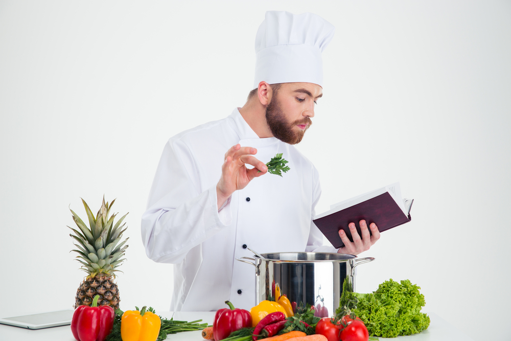 Portrait of a handsome male chef cook reading recipe book while preparing food isolated on a white background Portrait of a handsome male chef cook reading recipe book while preparing food isolated on a white background